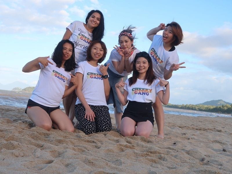 Experiencia en Australia grupo de 6 chicas posando en la arena y detrás el mar. Llevan la misma camiseta blanca con un estampado de letras en medio